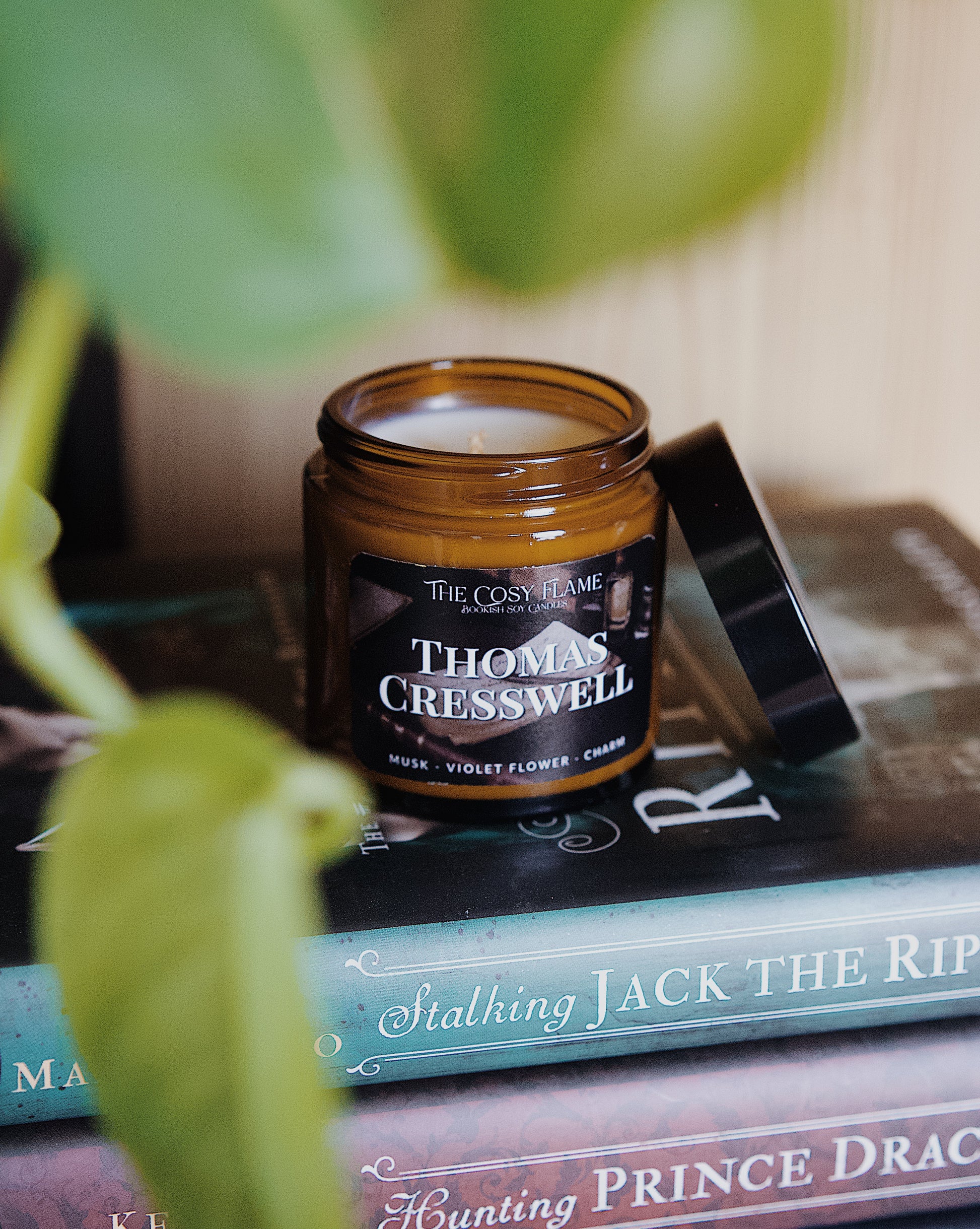 Amber glass candle jar labelled ‘Thomas Cresswell’ from The Cosy Flame, scented with musk and violet flower, and Stalking Jack the Ripper books in the background.