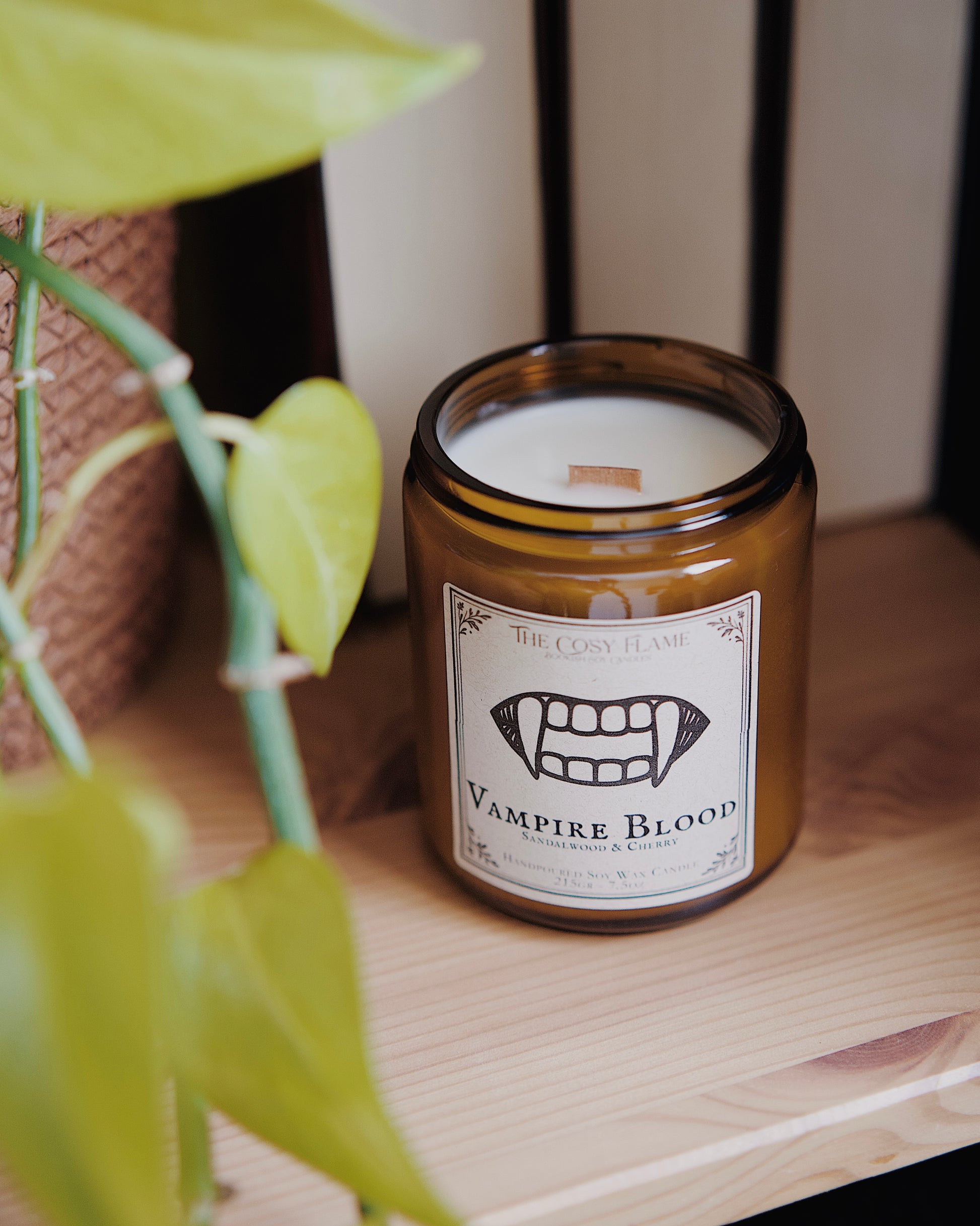 Vampire Blood soy candle by The Cosy Flame in an amber glass jar with a wooden wick, placed on a wooden shelf. Label features a vampire fangs illustration and featuring the scents sandalwood and cherry.
