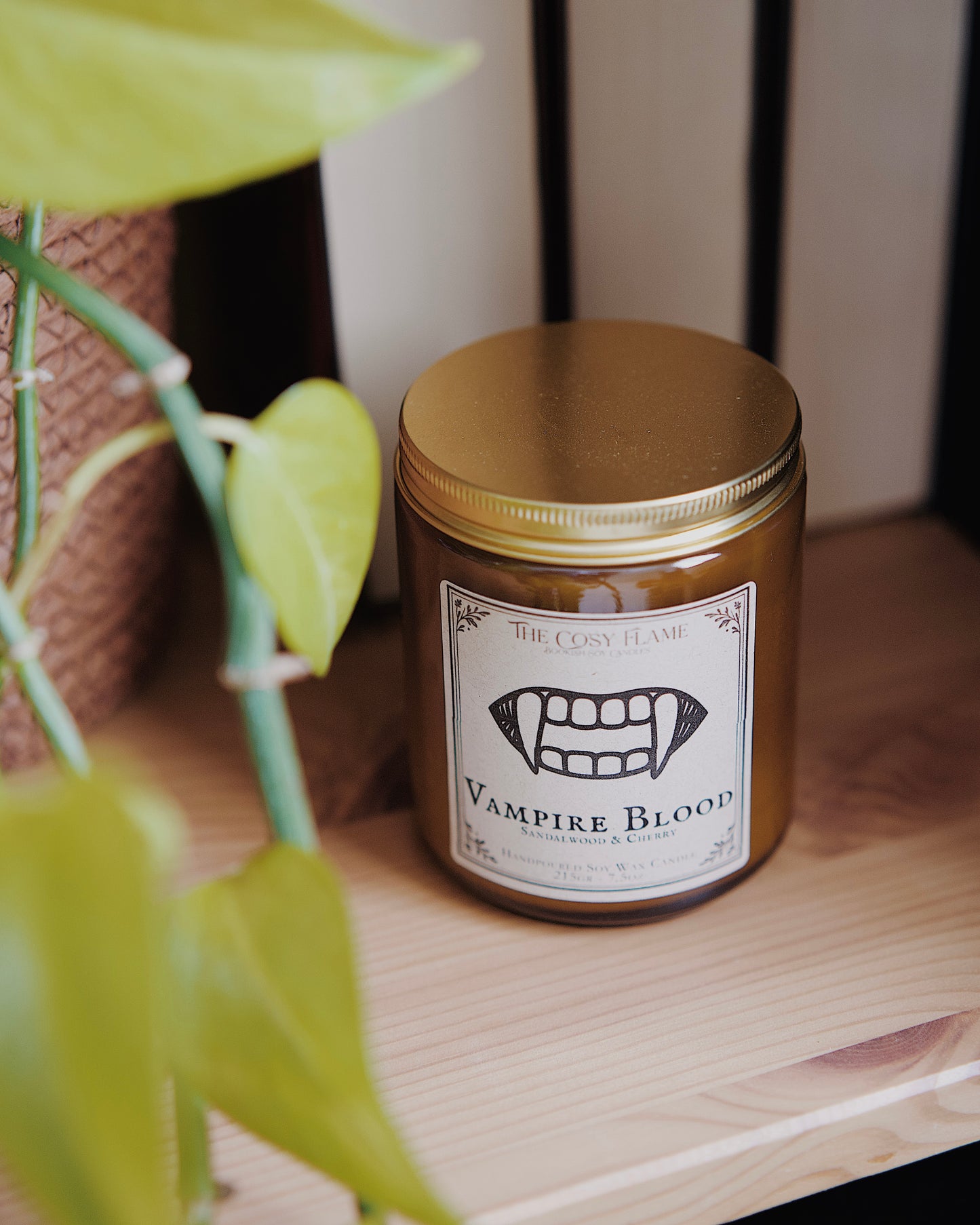 Vampire Blood soy candle by The Cosy Flame in an amber glass jar with a golden lid, placed on a wooden shelf. Label features a vampire fangs illustration and featuring the scents sandalwood and cherry.