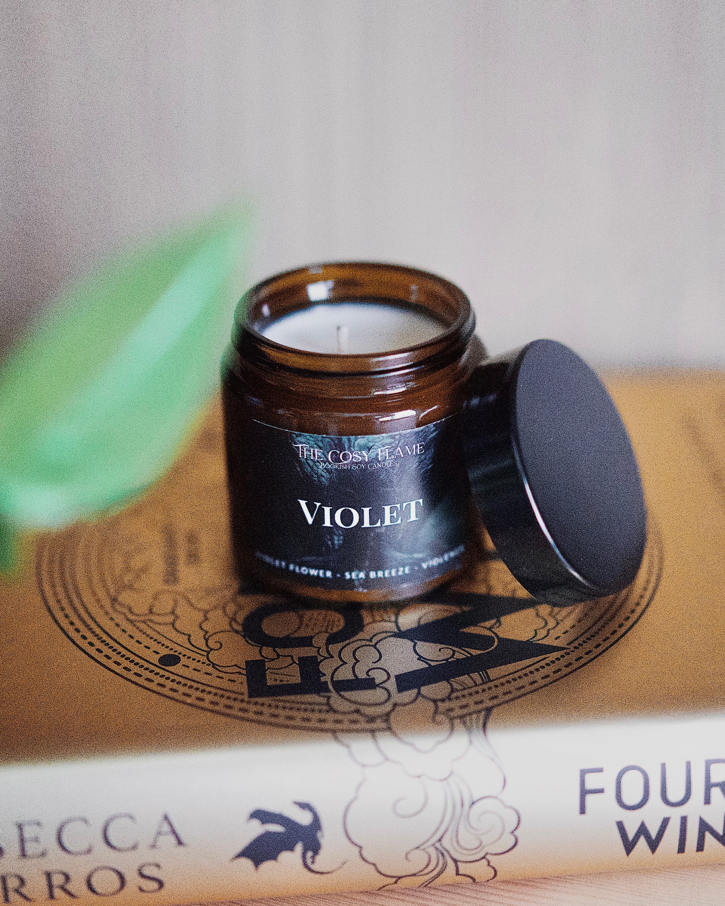 Amber glass candle jar labelled ‘Violet’ from The Cosy Flame, scented with violet flower and sea breeze, and Fourth Wing book in the background.