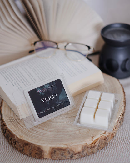 Violet soy wax melts by The Cosy Flame, featuring violet flower and sea breeze scent notes, displayed on a wooden slice beside an open book and glasses.