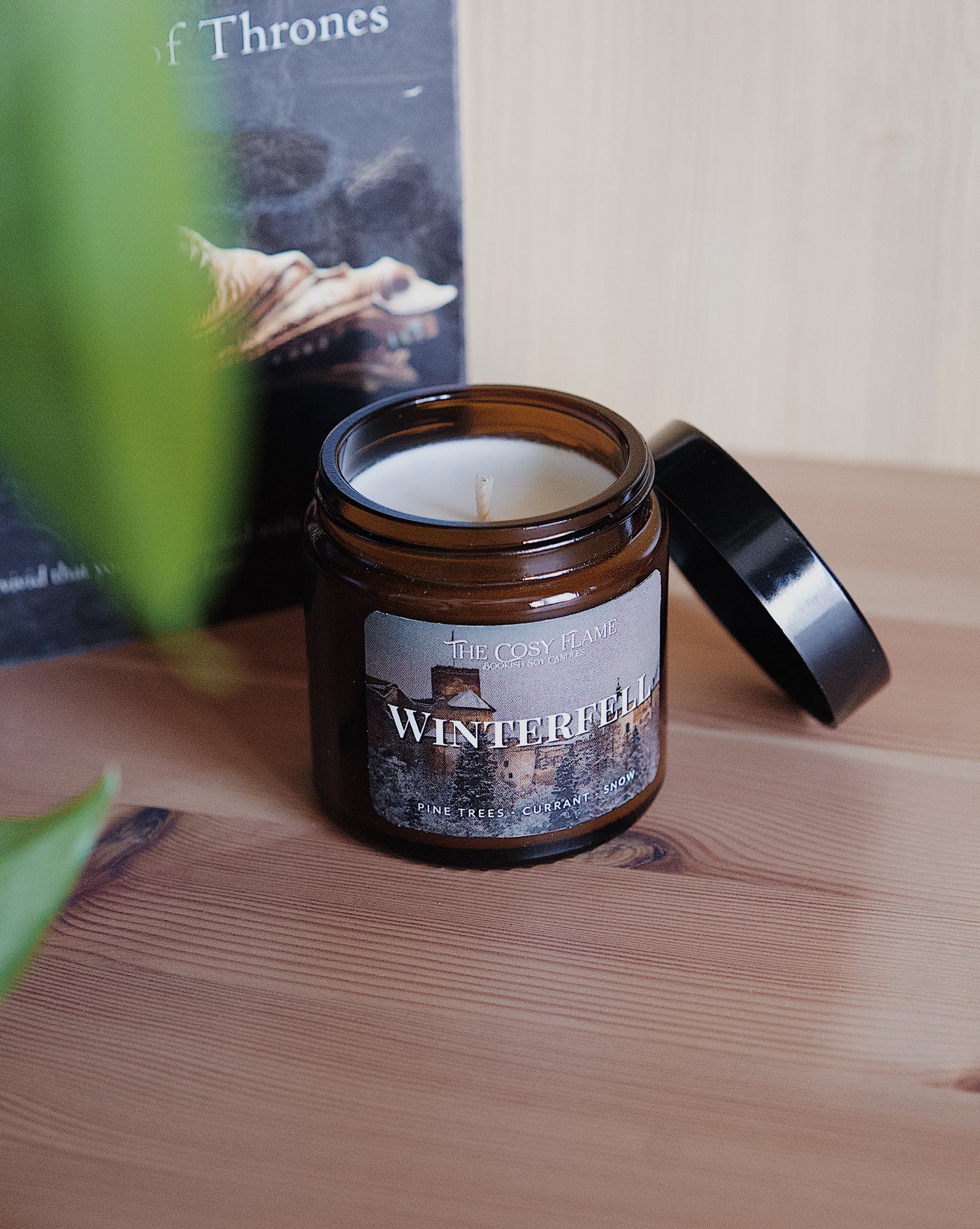 Amber glass candle jar labelled ‘Winterfell’ from The Cosy Flame, scented with pine trees and currant, and Game of Thrones book in the background.