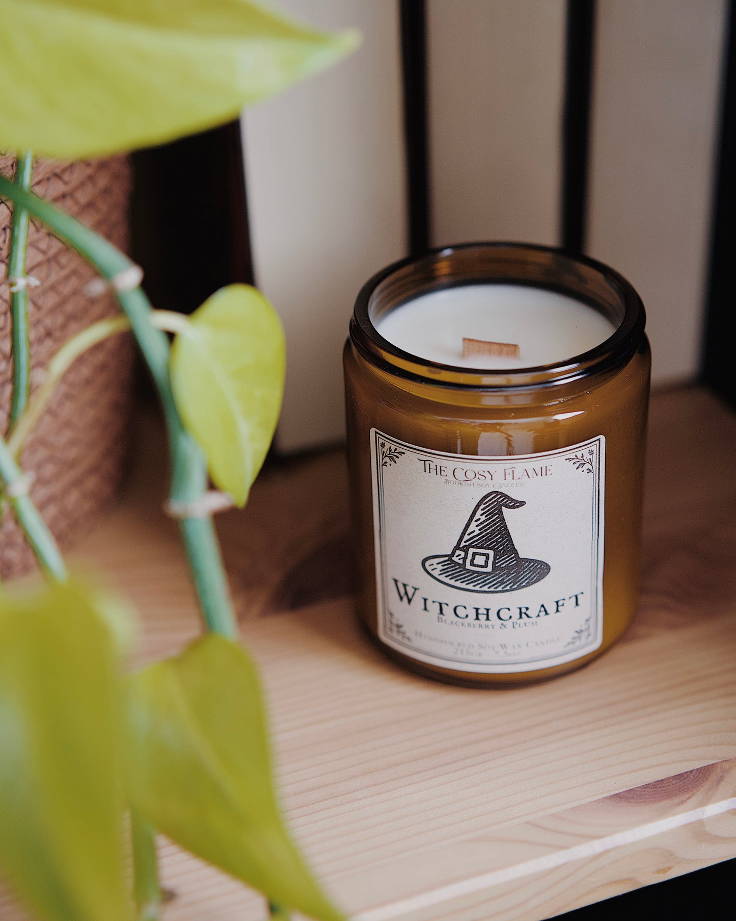 Witchcraft soy candle by The Cosy Flame in an amber glass jar with a wooden wick, placed on a wooden shelf. Label features a witch's hat illustration and featuring the scents
blackberry and plum.