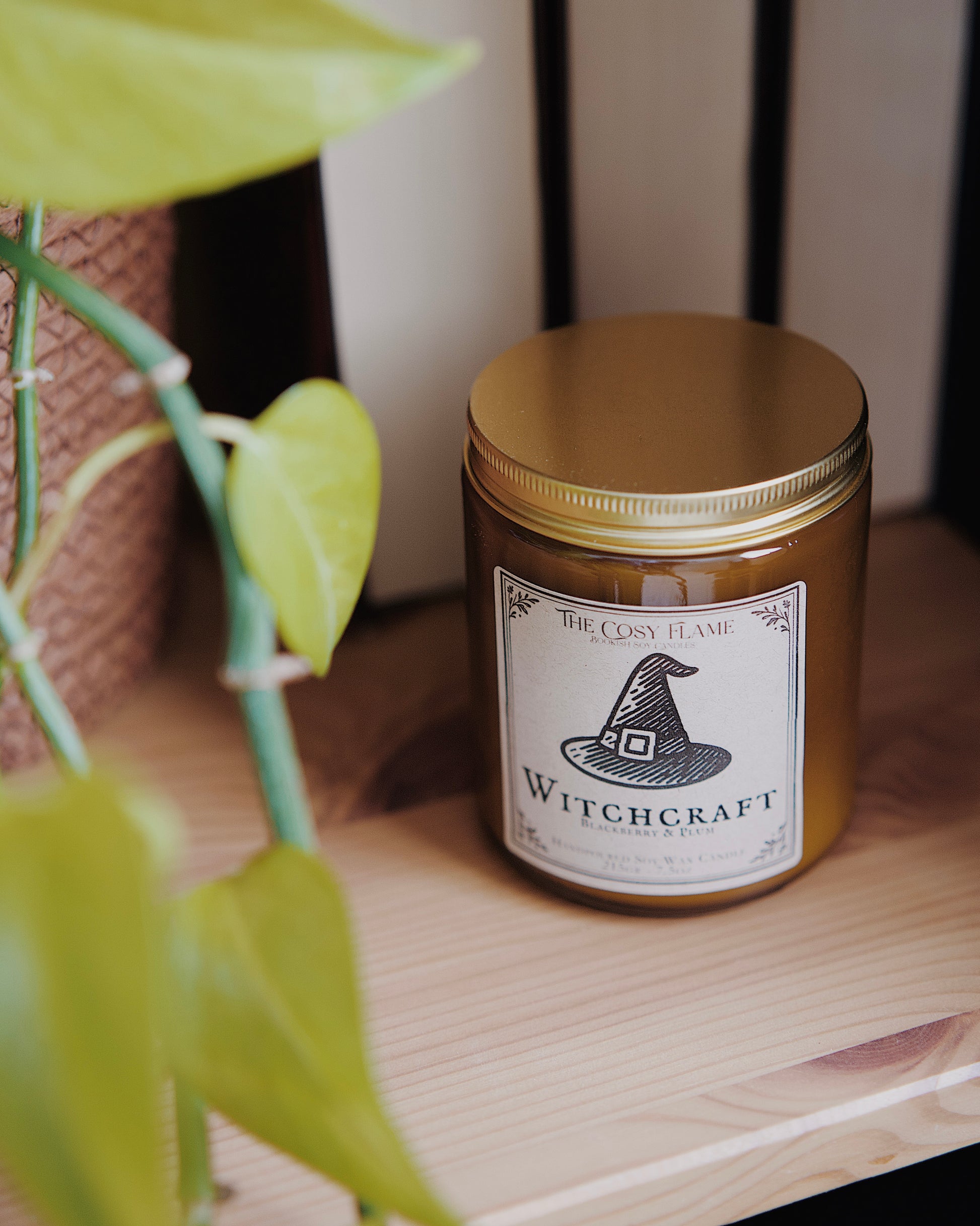 Witchcraft soy candle by The Cosy Flame in an amber glass jar with a golden lid, placed on a wooden shelf. Label features a witch's hat illustration and featuring the scents
blackberry and plum.